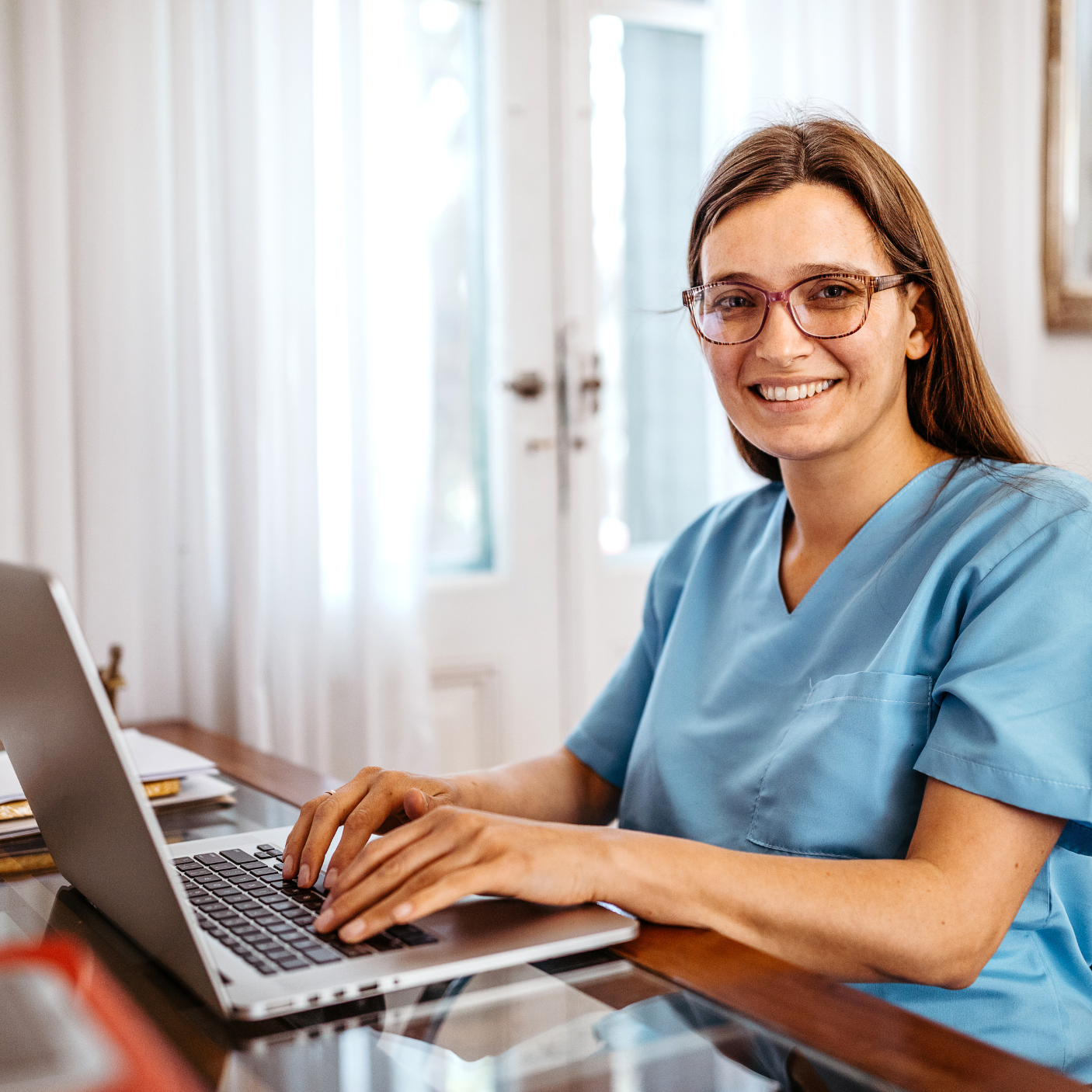 A white woman wearing blue scrubs sits at a desk. She wears glasses and smiles while using a laptop.