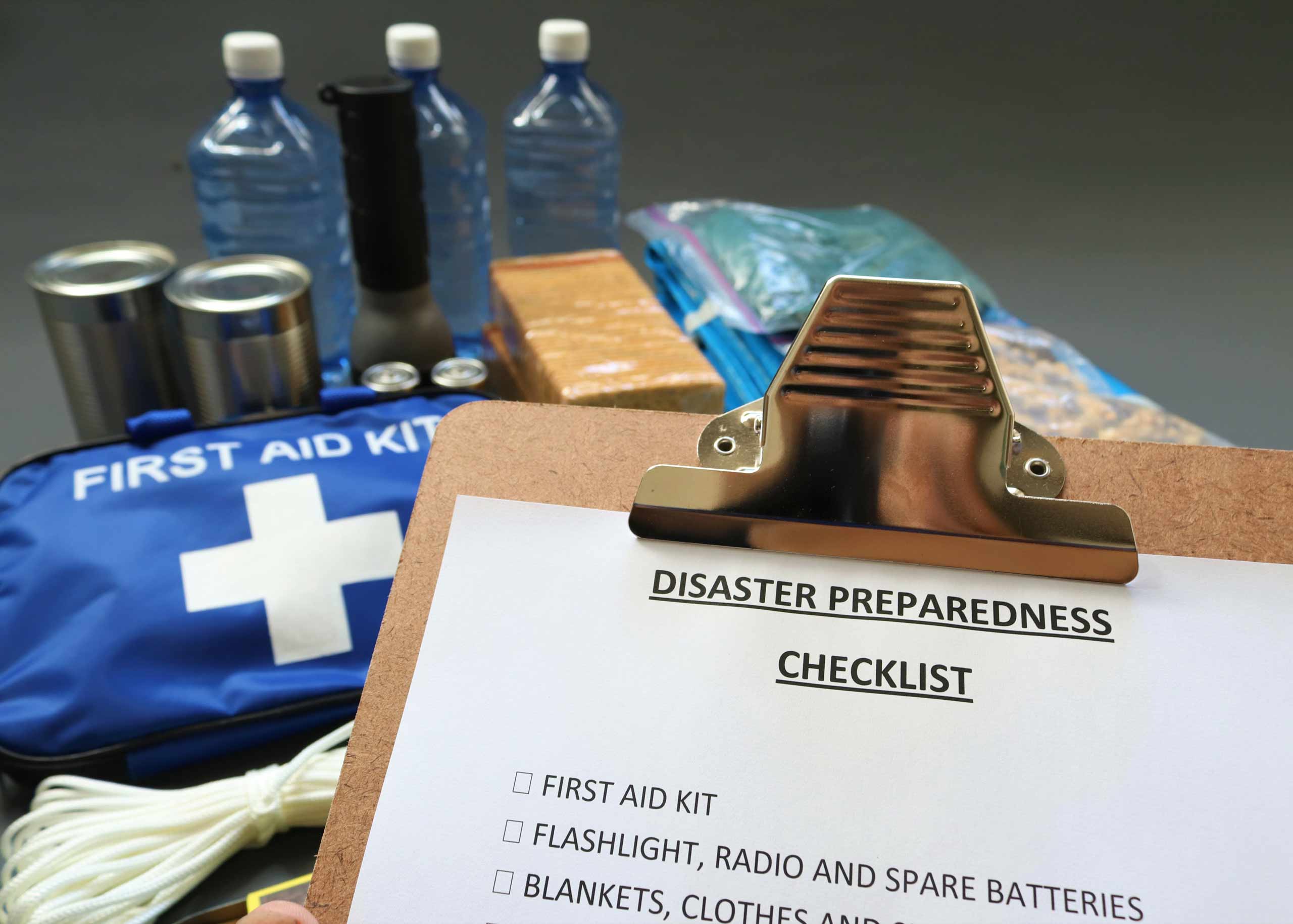 Disaster preparedness checklist on a clipboard with disaster relief items in the background.Such items would include a first aid kit,flashlight,tinned